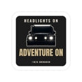 Headlights On / Adventure On die-cut sticker: a head-on 4x4 with glowing headlights between HEADLIGHTS ON and ADVENTURE ON lettering on black