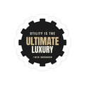 Utility Is the Ultimate Luxury die-cut sticker: UTILITY IS THE ULTIMATE LUXURY lettering in tan and white on a black gear-shaped badge